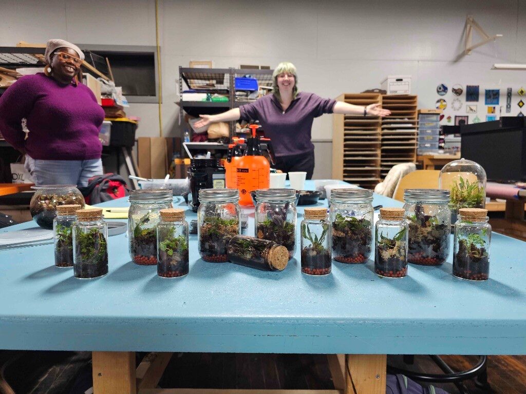 Table of handmade glass-jar terrariums with makers posing in the craft studio