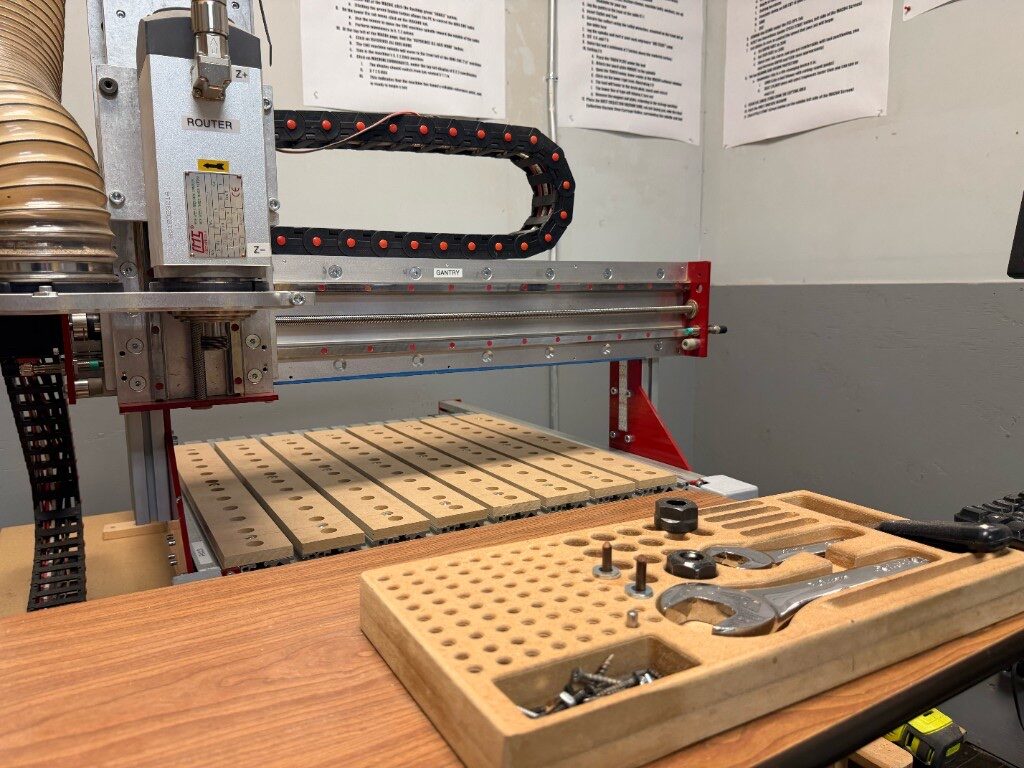 CNC router gantry and dust hose over a wooden spoilboard with a bit wrench tray in the foreground