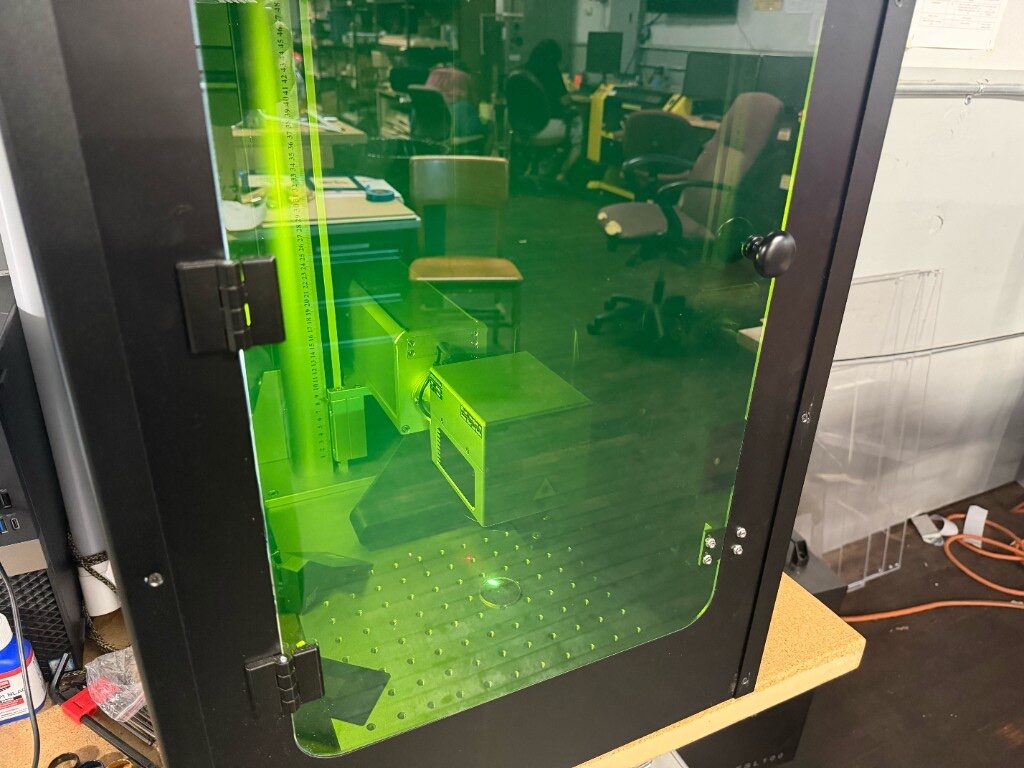 Laser engraver inside a green-tinted safety enclosure with the head over a perforated bed