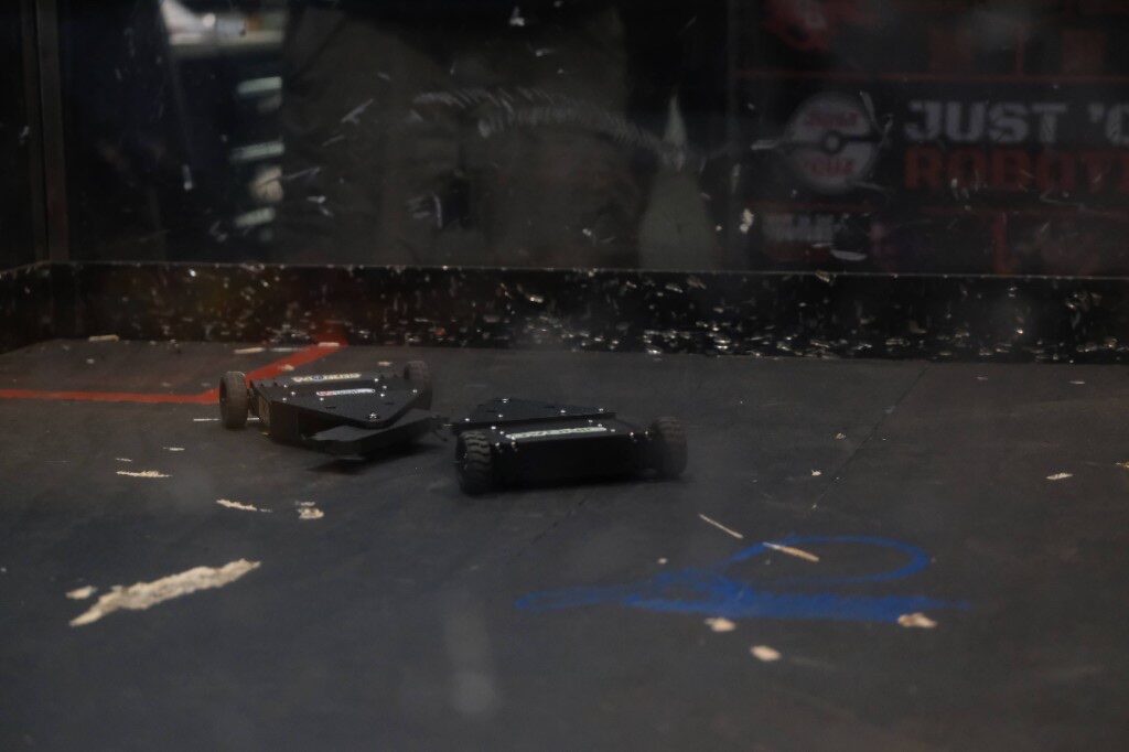 Two small black combat robots on a scratched arena floor behind a clear barrier